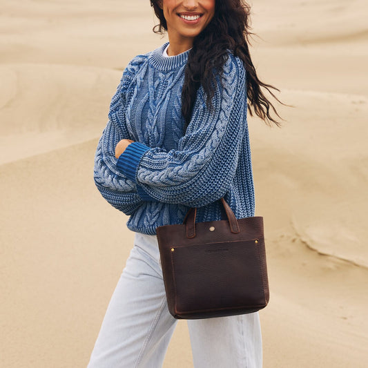 Model wearing blue knitted sweater carrying a leather Mini Crossbody Tote Bag in Coldbrew against a sandy beach bacground
