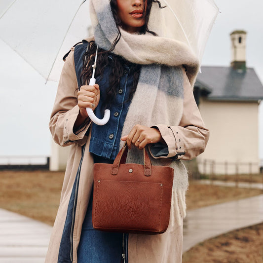 A model wearing a Mini Crossbody Tote in the color Nutmeg holding a clear umbrella