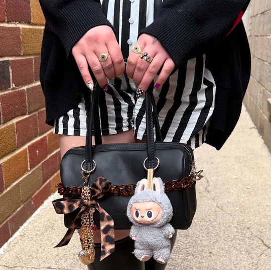 A pair of hands holding a black leather handbag decorated with a printed bow, gold chain, and a grey labubu stuffed animal keychain.