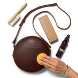 4oz | Hand applying Natural Leather Care to a circle bag with a sponge surrounded by a brush and the bottle and box.