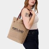 Woman holding a canvas tote bag with 'Portland Leather Goods' branding.