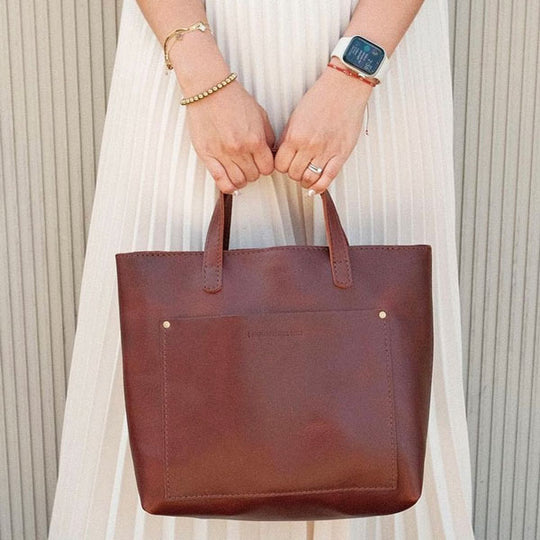 Brown leather tote bag held by a person against a neutral dress and wall