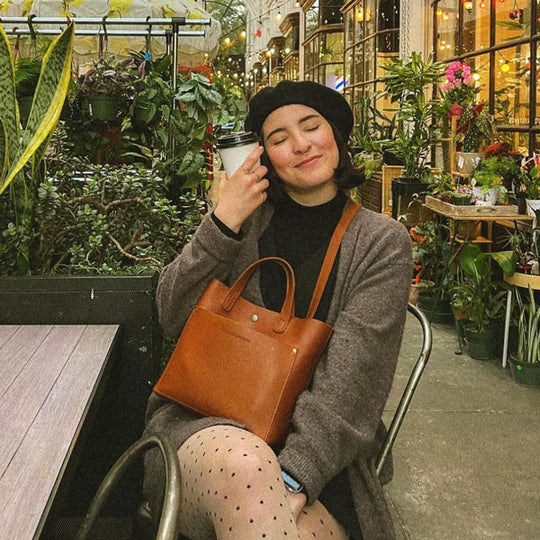 Woman sitting in a greenhouse holding a coffee cup with a brown leather bag on lap
