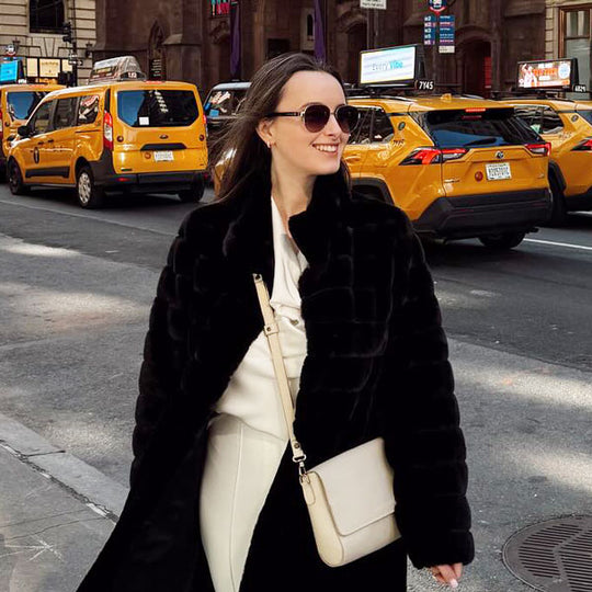 Woman in a black coat sunglasses and a white crossbody bag standing on a city street with yellow taxis in the background
