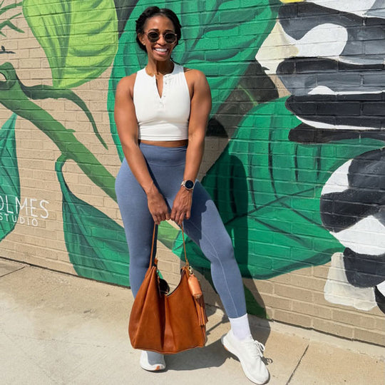 Woman in athletic wear standing in front of a colorful mural holding a brown purse