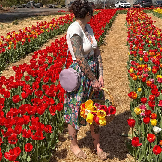 Woman standing among rows of tulips holding a basket, with a field of tulips in the background wearing a crossbody bag