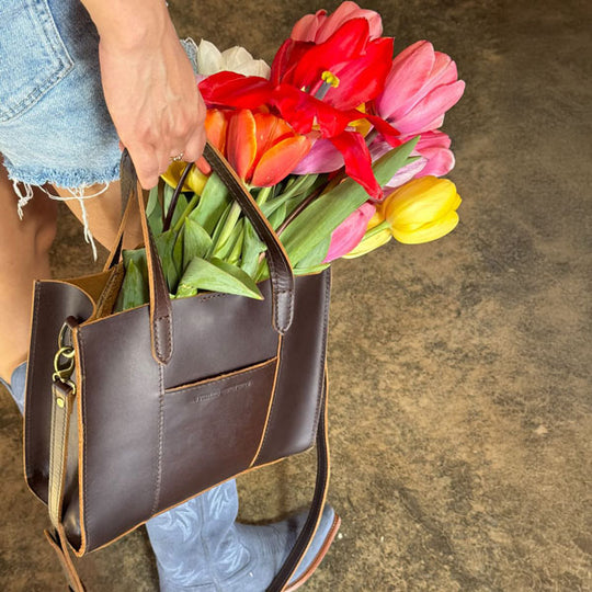 Person holding a brown leather handbag filled with colorful tulips on a textured surface.