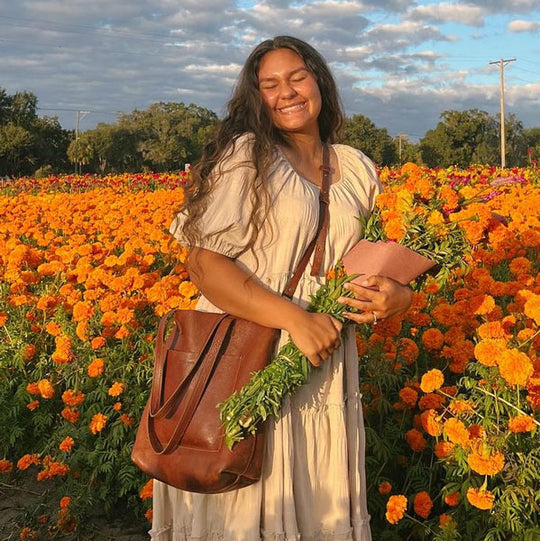 Woman standing in a field of marigolds holding a bouquet and a brown bag.