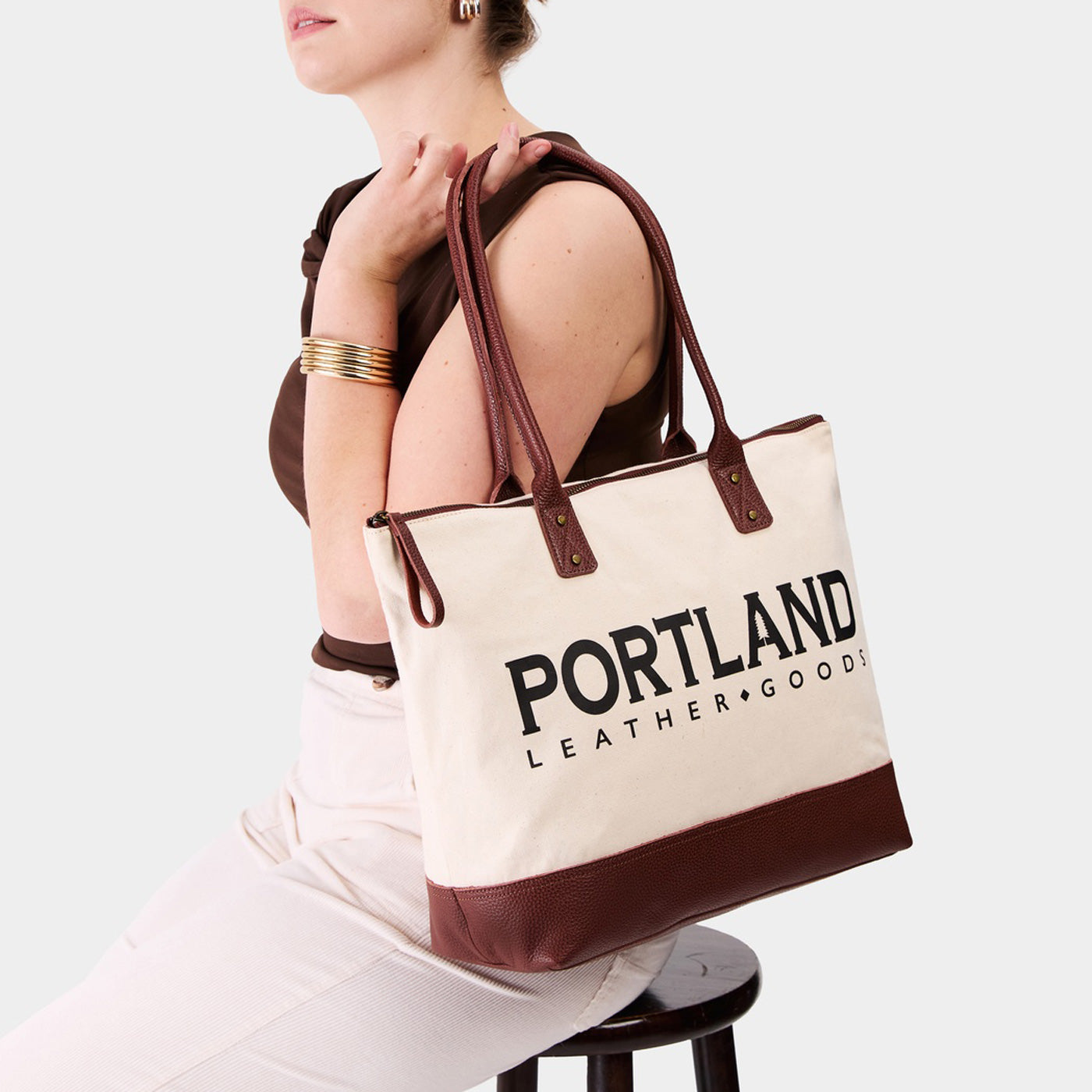 Model holding canvas tote with zipper, leather bottom, and leather handles on a white background