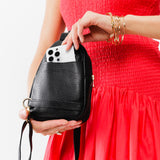 Pebbled--black | Model putting phone into pocket on the back of a small sling bag with zipper closure and adjustable strap
