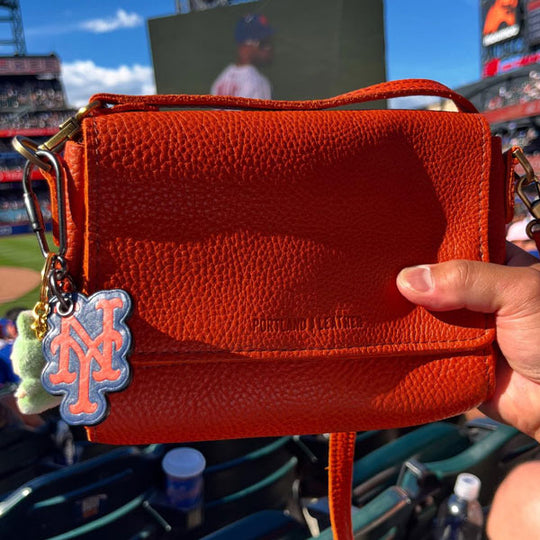 Hand holding a rectangular orange leather bag with a New York Yankees keychain. Baseball stadium in the background.