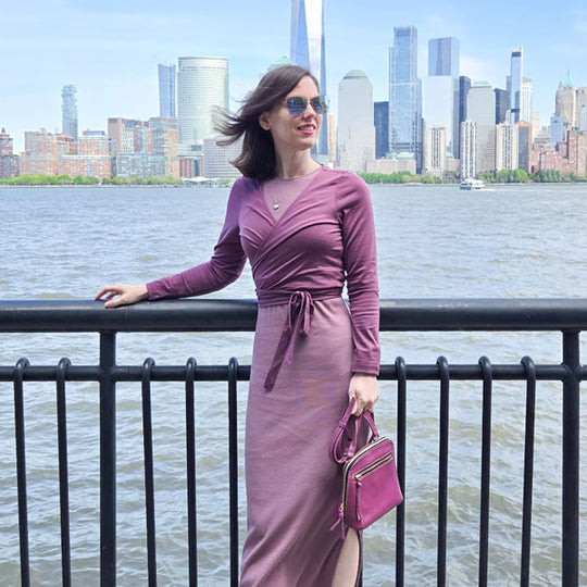 Woman in a purple dress with a purple leather purse standing in front of a metal railing with the New York city skyline and the bay in the background.