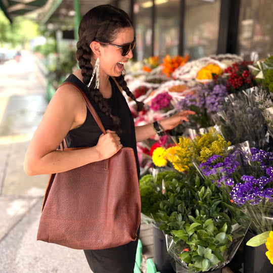 Woman in sunglasses and braids carrying a brown rectangular leather tote bag in front of a flower shop with a storefront full of flower bouquets.