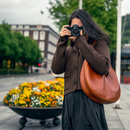 Woman holding up a camera to her face and carrying a brown crescent-shaped leather shoulder bag in front of a city street with flowers in a large planter.