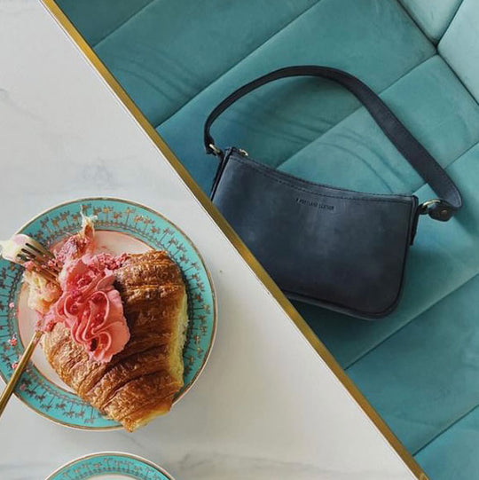 Croissant on a plate on table with a deep blue leather shoulder bag laying flat on the padded bench next to it.