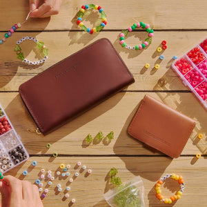 Two embossed leather zip-around wallets, shown in a styled flat-lay on a wooden table surrounded by colorful beaded bracelets, beads, and small accessories. One wallet is dark brown and the other is tan.